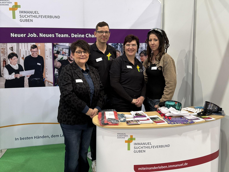 Marion Swietza, Doreen Lindner, Steffi Mattke und Christopher Betz vom Immanuel Suchthilfeverbund Guben stehen an dem mit Logos und Bannern gestalteten Messestand vor einem Tisch mit Informationsmaterialen und Giveaways auf der Ausbildungsmesse Impuls in Cottbus und blicken in die Kamera 