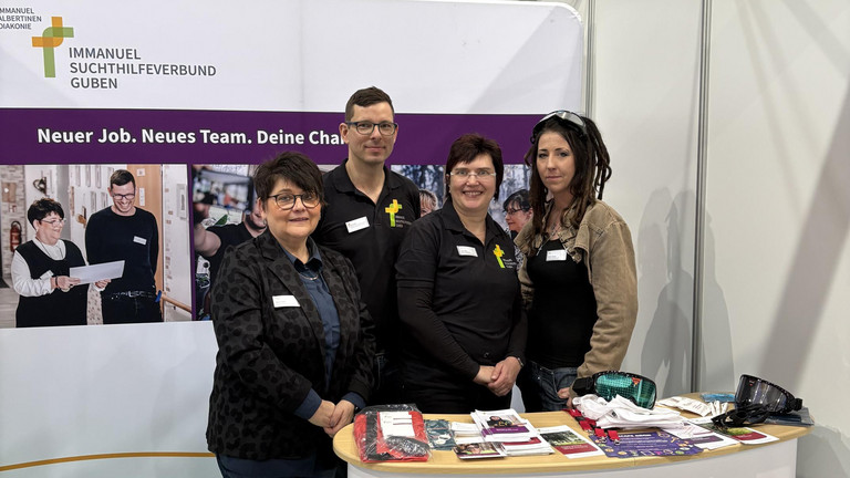 Marion Swietza, Doreen Lindner, Steffi Mattke und Christopher Betz vom Immanuel Suchthilfeverbund Guben stehen an dem mit Logos und Bannern gestalteten Messestand vor einem Tisch mit Informationsmaterialen und Giveaways auf der Ausbildungsmesse Impuls in Cottbus und blicken in die Kamera 